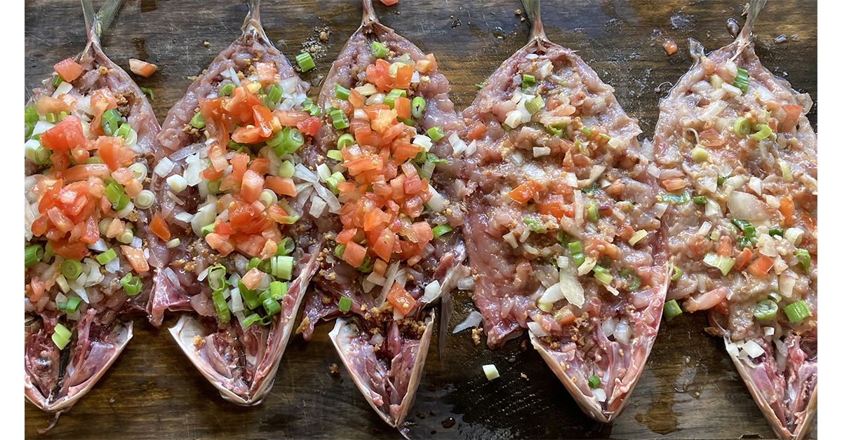 Image of five ʻōpelu fish lined up in stages of preparation with chopped onions, tomatoes, seaweed, and kukui nut.